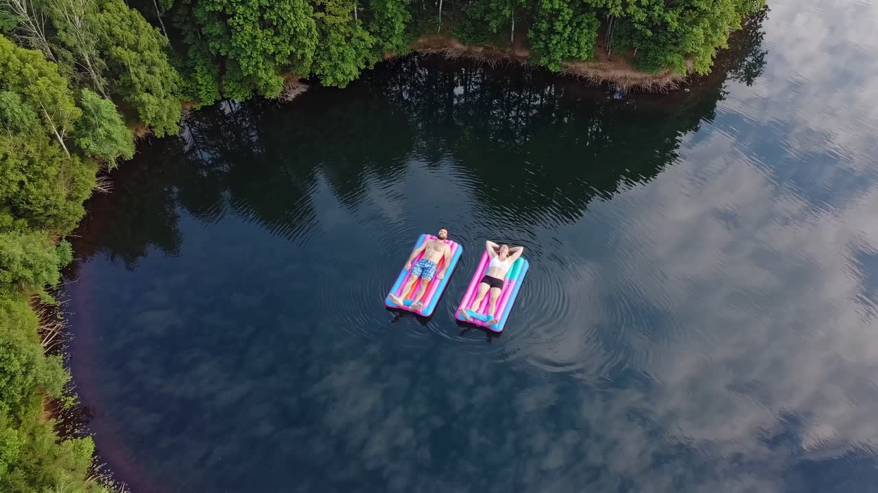 Aerial view of a couple sunbathing and relaxing on colorful inflatable mattresses in a calm lake surrounded by lush green forest, enjoying a peaceful summer day