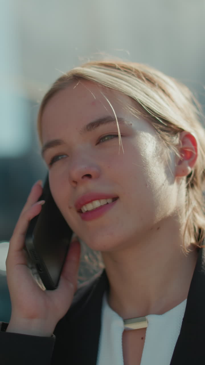 Young lady in professional attire with warm smile on phone call walking by glass building under bright sunlight, city reflections visible on glass, radiating positivity and calmness in urban environment