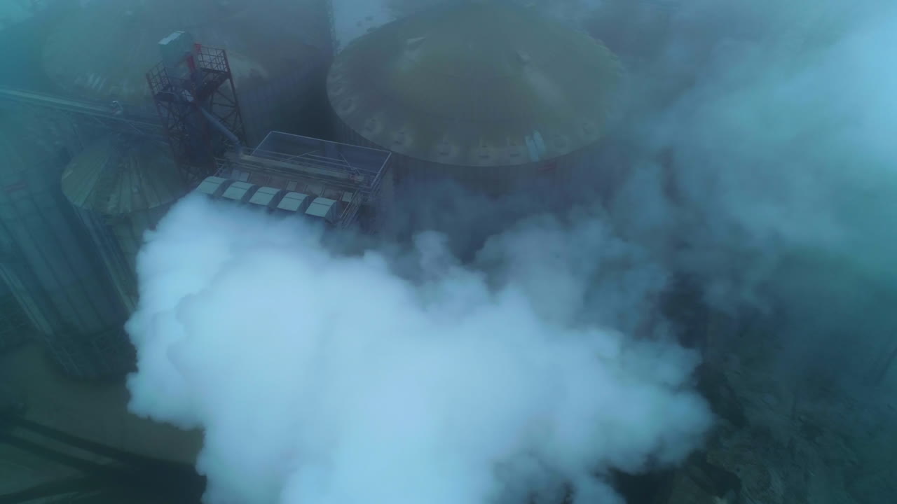 Huge functioning agribusiness plant for grain storage and processing. Top view on the silo tanks roofs in the white smoke and fog.