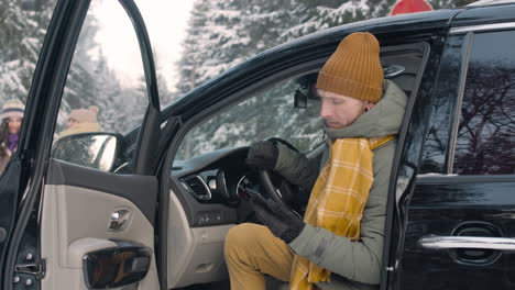 Mann-In-Winterkleidung,-Der-Im-Smartphone-Sitzt-Und-Mit-Offener-Tür-In-Einem-Verschneiten-Wald-Im-Auto-Sitzt