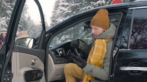 mann in winterkleidung, der im smartphone sitzt und mit offener tür in einem verschneiten wald im auto sitzt