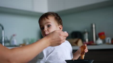 adorable caucasian toddler sitting at the table and holding a piece of bread. mom fills the spoon in the bowl but kid refuses to eat.
