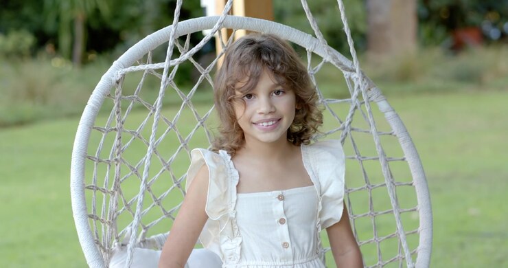 La cámara se amplía, niña con vestido color crema cambiando de postura y sonriendo en una silla de jardín tejida.