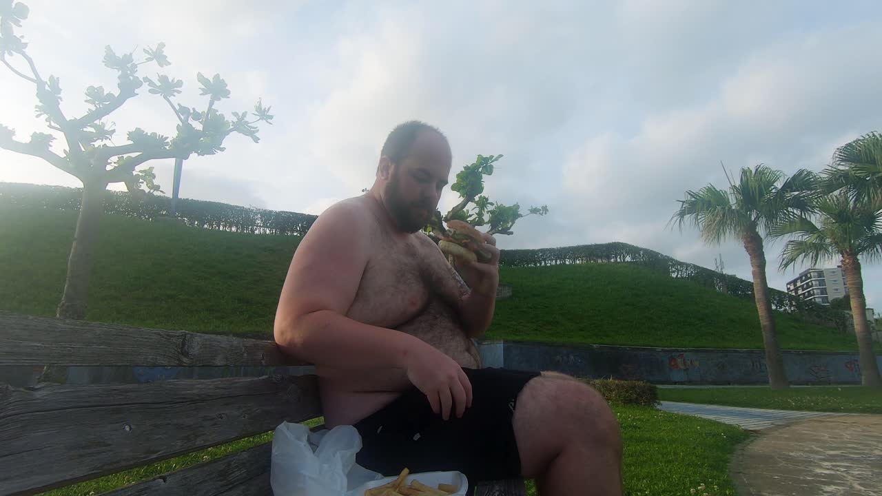 공공 공원 벤치에 앉아 뚱뚱하고 못생긴 배를 드러내면서 기름진 햄버거를 먹는 뚱뚱한 남자