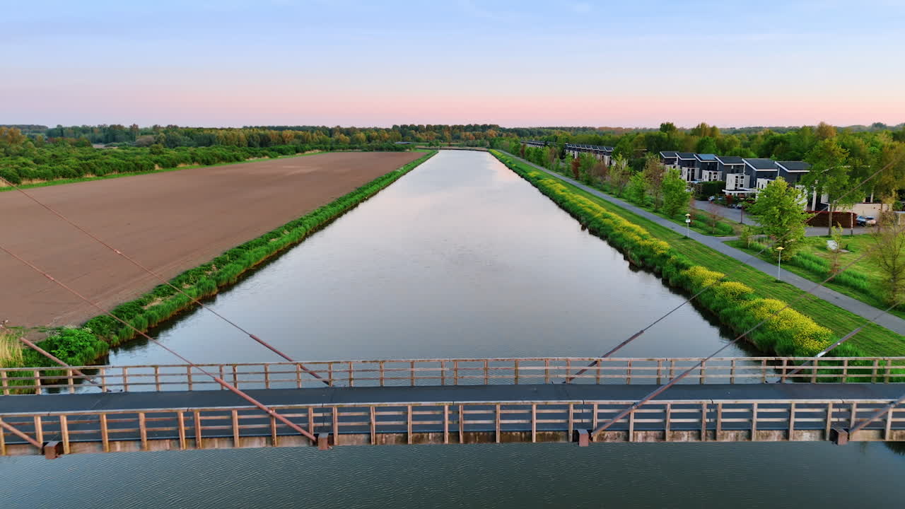 Bridge over calm water, Netherlands. Sunset casts a warm glow on the bridge spanning a serene canal, surrounded by lush greenery and fields