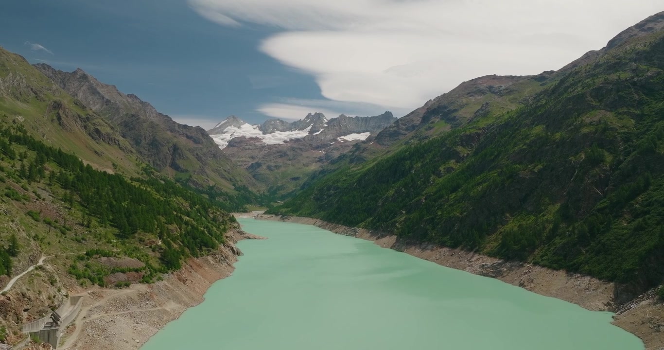 Il drone si avvicina a un'imponente diga con vivida acqua turchese del bacino dietro di essa, circondata da cime alpine innevate sotto un cielo estivo azzurro e limpido