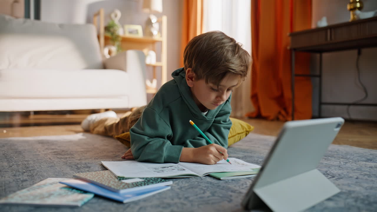 Diligent schoolboy writing exercise notebook looking tablet lying home floor