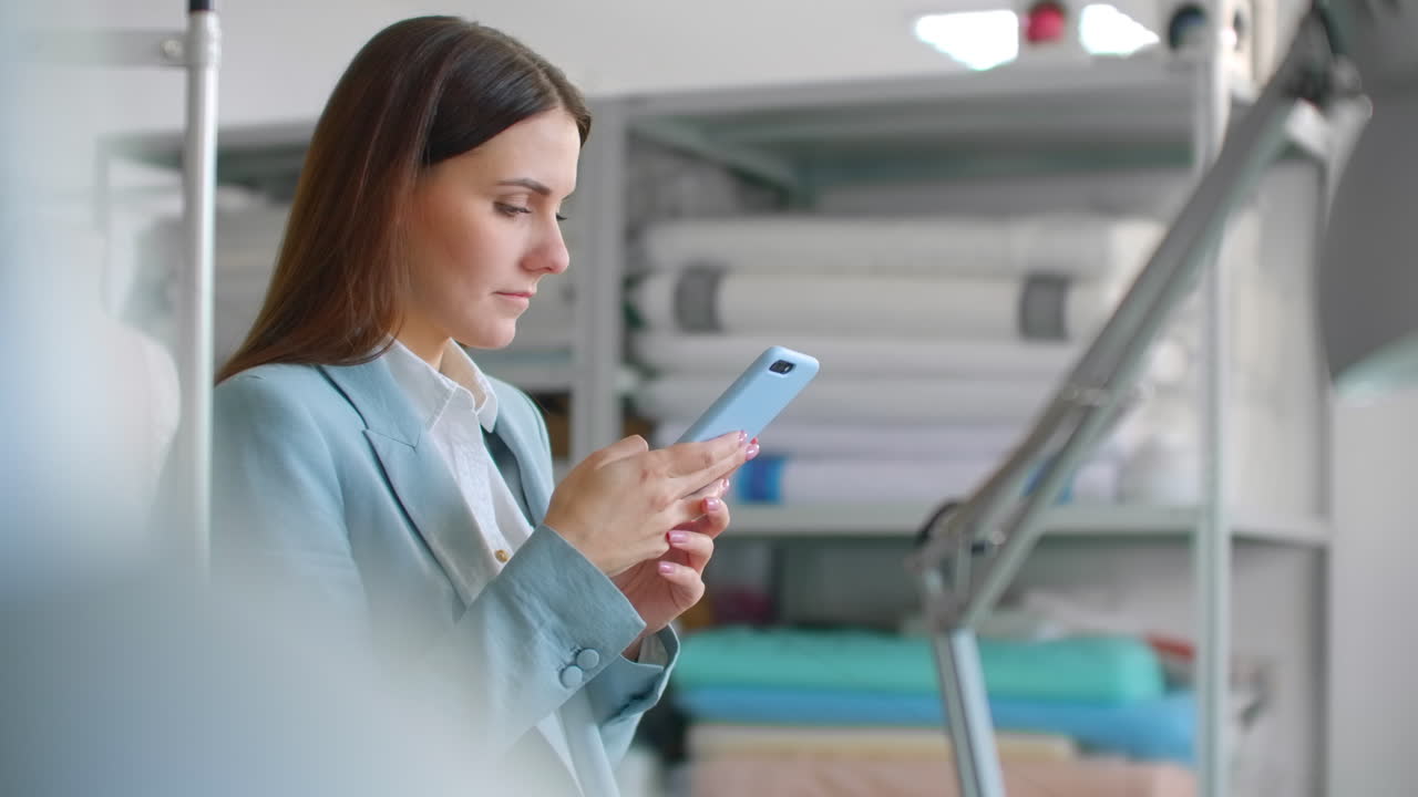 joven mujer diseñadora mujer de negocios en el estudio de costura con teléfono móvil en el taller de costura escribiendo mensajes a los clientes