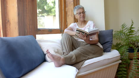 Happy-senior-biracial-woman-on-couch-reading-book-at-window-at-home,-slow-motion