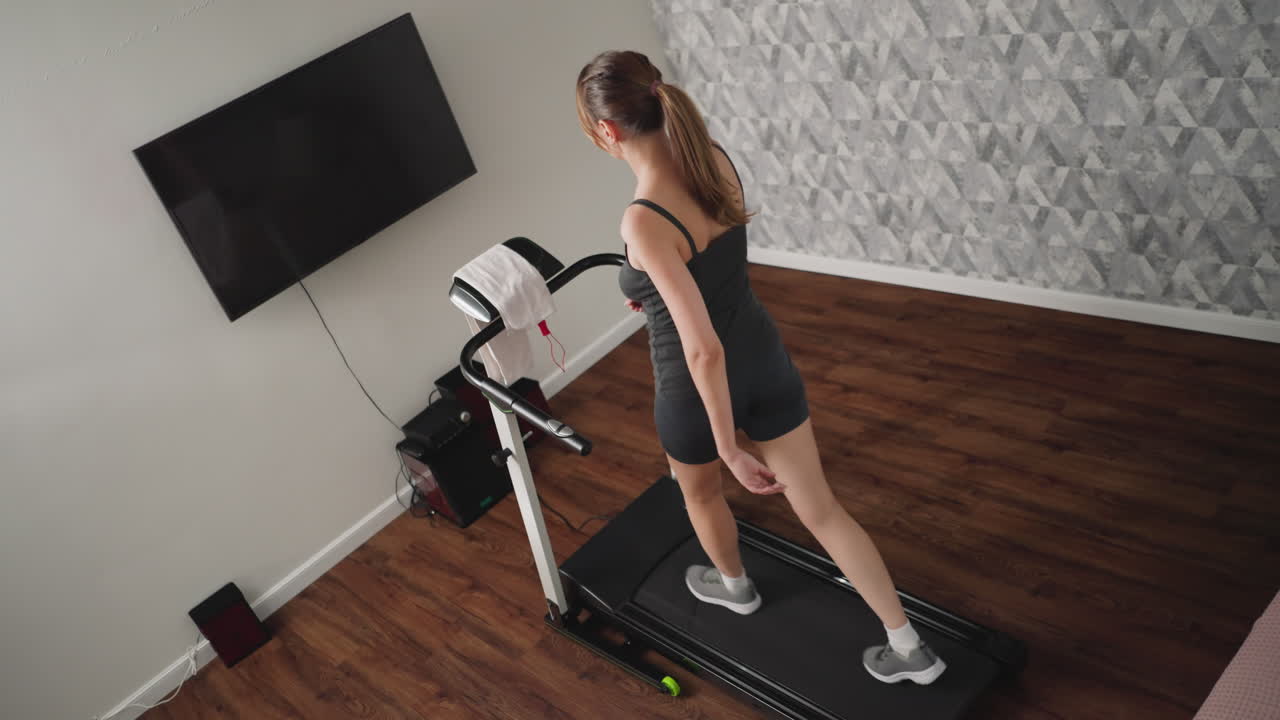 Rear view of woman with ponytail walking on treadmill in cozy indoor space with wooden floor, towel draped over console, wall-mounted TV and sound system in background