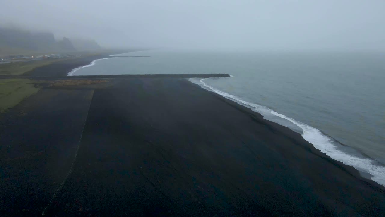 Drone soars over Vik's black sand beach, where fog enshrouds crashing waves, unveiling a hauntingly beautiful scene in sharp 4K detail