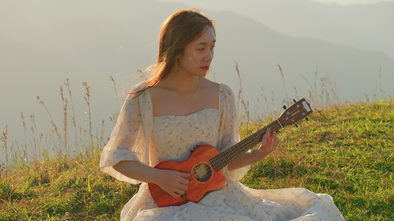 asiatic woman artist musician playing ukulele guitar in nature at sunset sitting on green meadow wearing long white dress
