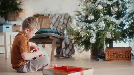 little boy opens christmas present in front of christmas decorations and christmas tree is happy