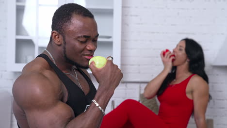 multi ethnic couple with apples in the kitchen. muscular african man eating apple on the background of a sexy woman in red dress sitting on the kitchen table.