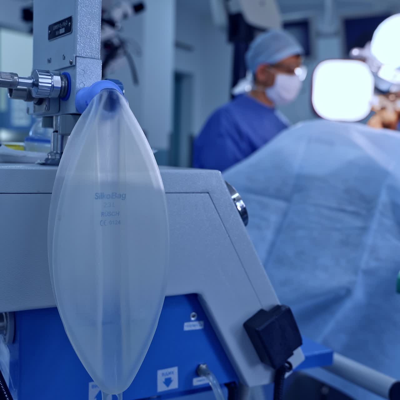 Slightly inflating ambu bag hanging on the technological equipment. Medical team performing surgery at backdrop in blur