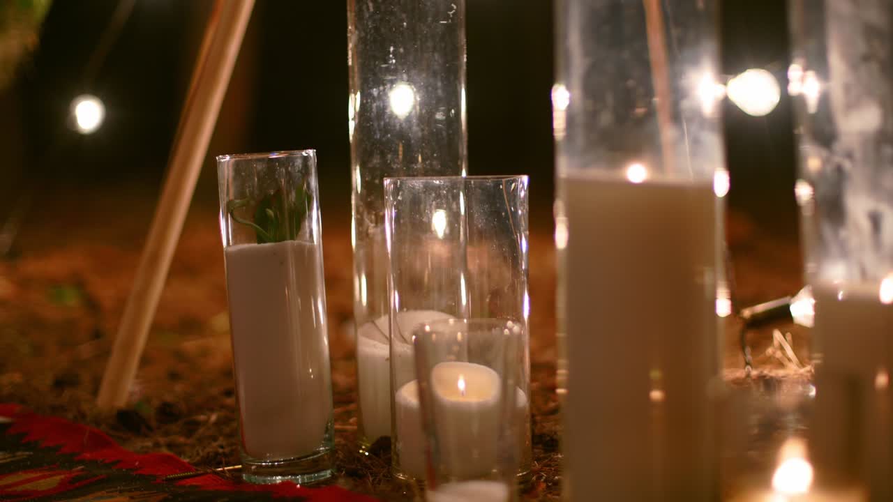 Candles burning in glass vases, flasks on carpet near bohemian tipi arch decorated with roses and flowers wrapped in fairy lights on outdoor wedding ceremony venue in pine forest. Boho rustic decor