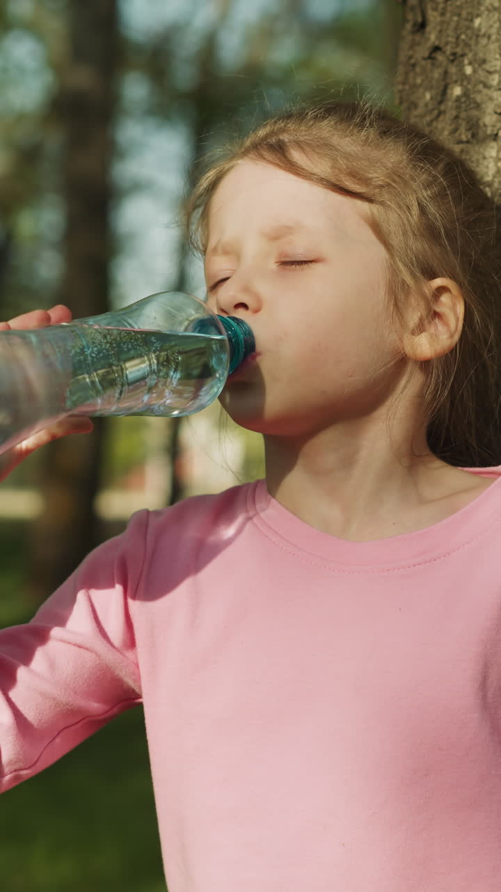 Thirsty blonde girl drinks clear water near old tree in city park closeup. Refreshing beverage for child in hot weather. Health care and wellbeing