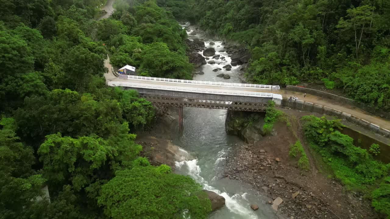 Aerial view of a concrete-and-metal bridge crossing a rocky river in a lush Philippine forest, with white railings, misty hills, and winding roads blending nature and engineering