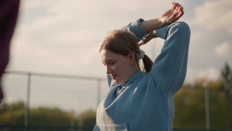 young athlete in blue sweater exercising outdoors with sunlight highlighting her face, with a partial view of someone nearby in a sports arena