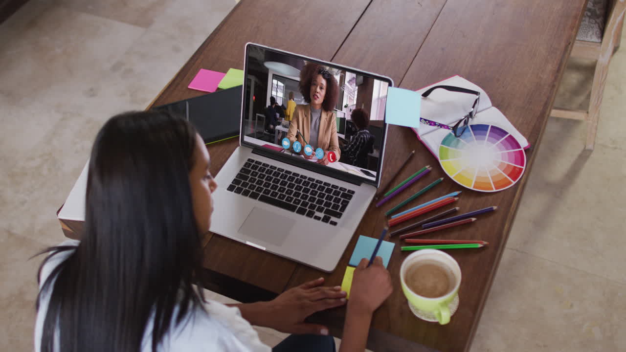 Mujer afroamericana escribiendo en notas adhesivas mientras realiza una videollamada en su computadora portátil en casa.