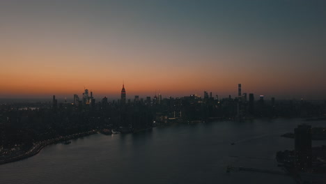 AERIAL:-Over-East-River-showing-Manhattan-New-York-City-Skyline-in-Beautiful-Dawn-Sunset-Orange-Light-right-before-Dark