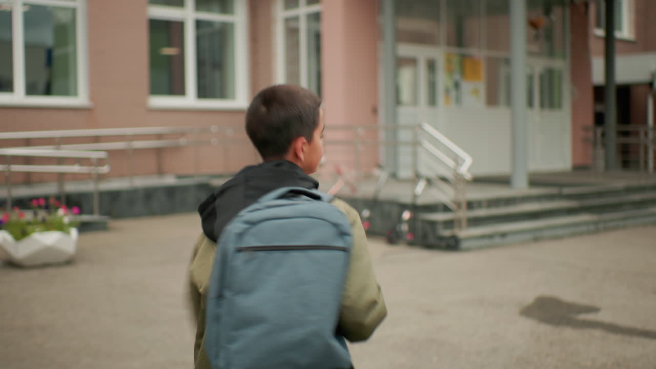 Vista trasera de un estudiante con mochila corriendo hacia la entrada de la escuela; se ven las marcas en el suelo y los escalones, con el exterior del edificio al fondo.