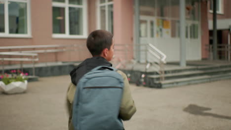 back view student with backpack running toward school entrance, visible ground markings and steps ahead, building exterior in background