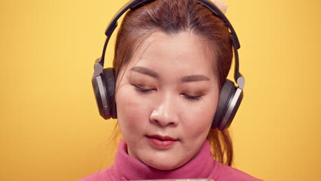 밝은 노란색 배경에서 편안한 헤드폰으로 음악을 즐겁게 듣기 위해 디지털 태블릿과 스트리밍 응용 프로그램을 사용하는 아름다운 아시아의 화려한 젊은 여성