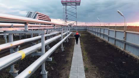 a drone flies after a worker in overalls and a helmet, who walks along a path along industrial pipes. extraction of oil and gas in industry. oil refining