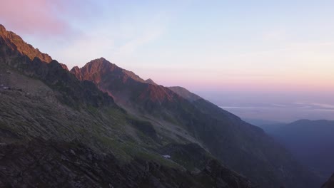 Stunning-aerial-panoramic-view-of-jagged-mountain-peaks-of-Romania-and-rugged-terrain-with-colorful-light-of-sunset-on-the-landscape-and-distant-clouds