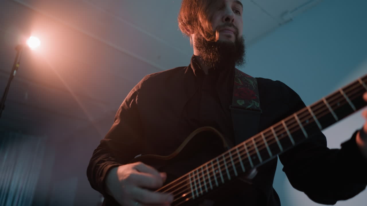 A guitarist in a black outfit, standing and playing a red electric guitar against a blue background with a bright light in the background. The musician appears deeply focused on the performance