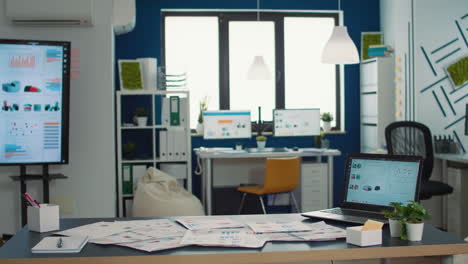 Empty-startup-office-with-laptop-showing-analytics-on-display-with-moving-charts-and-tv-wall-screen