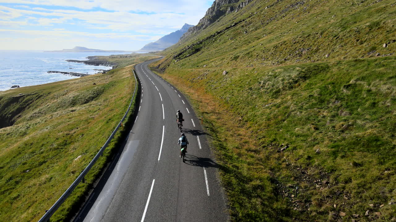 Drone Following People Biking Around Bend in Road On Iceland Road Over Ocean Toward Mountains