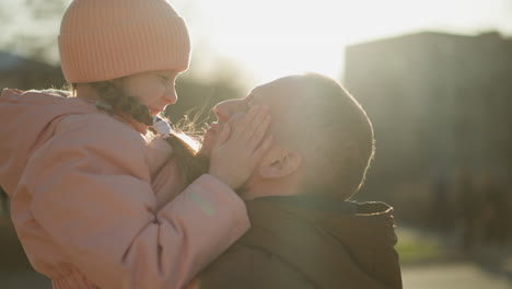 a joyful little girl in a pink cap and jacket is being carried by a man in a brown jacket. she is playfully touching his face and hugging him tightly, both smiling warmly as they share a tender moment