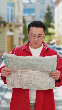 asian man tourist exploring city street sightseeing holding paper map check direction searching way