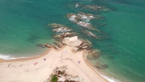 Slow-Drone-video-over-sandy-summer-beach-rock-formation-waves-people-relaxing