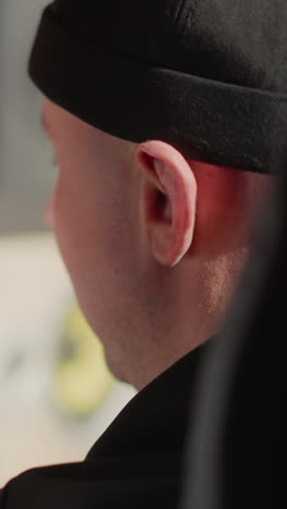 toma lateral de primer plano de un hombre con una gorra negra, con su oreja prominentemente visible. un micrófono está en el fondo borroso, lo que sugiere una configuración de grabación o producción de audio