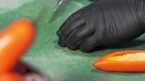 chef, con un delantal de denim azul y guantes estériles negros, cortando y cortando hábilmente pimientos de chile naranja peruanos frescos en tiras en una tabla de corte de plástico verde usando un cuchillo de chef afilado