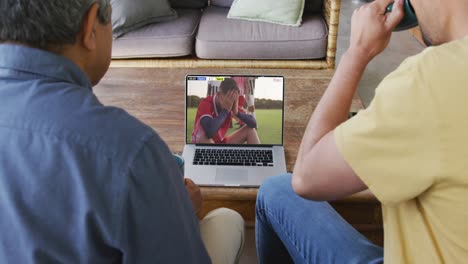 biracial father and son watching laptop with diverse male soccer players playing match on screen