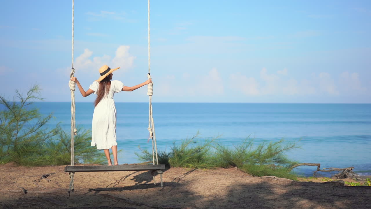 de vuelta a la cámara, una mujer joven con un vestido de verano y un sombrero de paja se para en un gran columpio de madera con vista al horizonte del océano