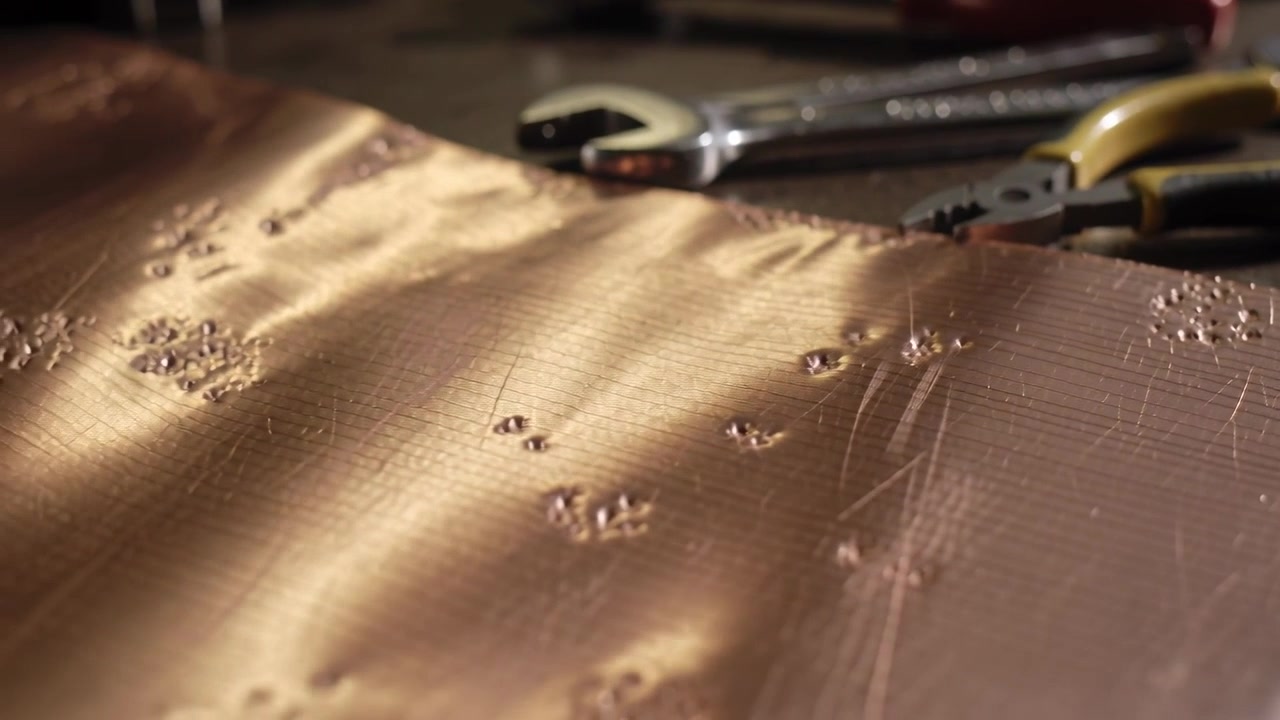 Moving camera sliding right and closer across bench, revealing copper sheet ripples, pliers, wrench