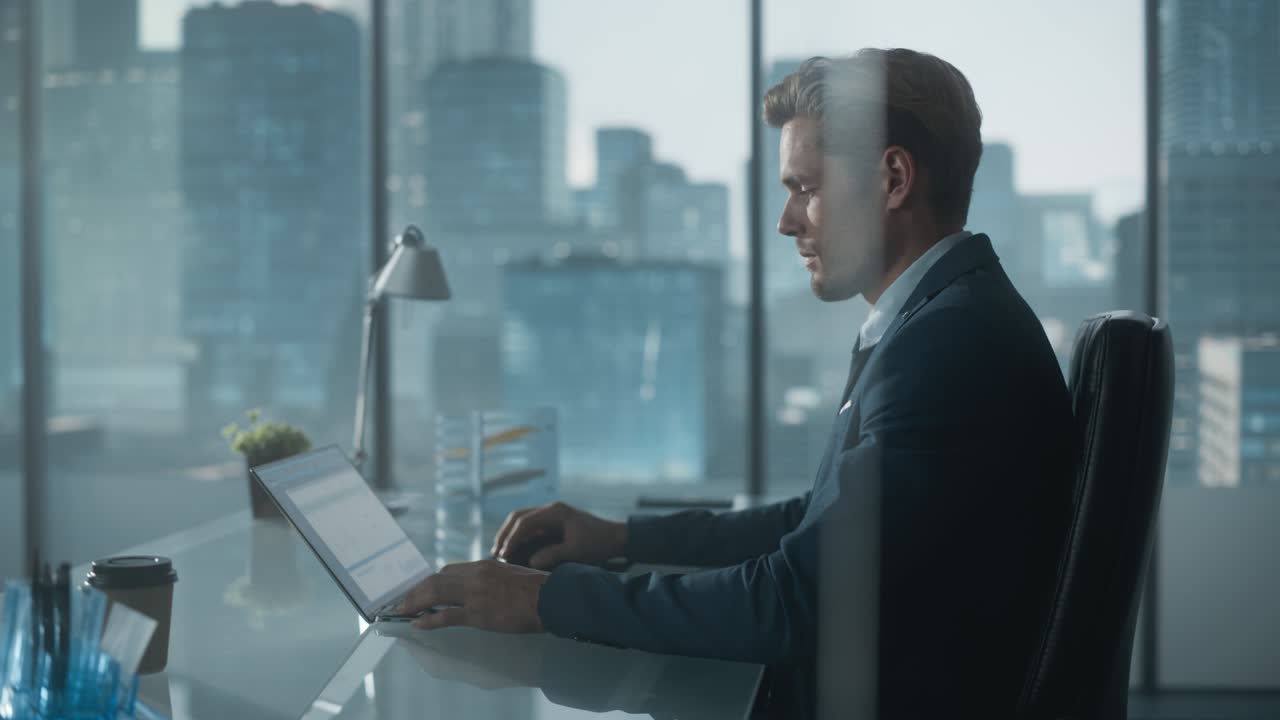 Confident Businessman in a Suit Sitting at a Desk in Modern Office, Using Laptop Computer, Next to Window with Big City with Skyscrapers View. Successful Finance Manager Planning Work Projects.