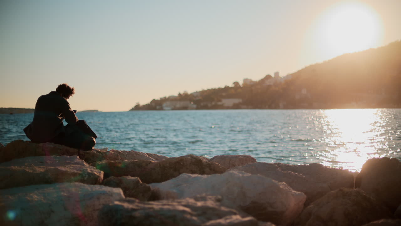 A man sitting alone on large rocks by the sea, looking toward the horizon under a clear sky