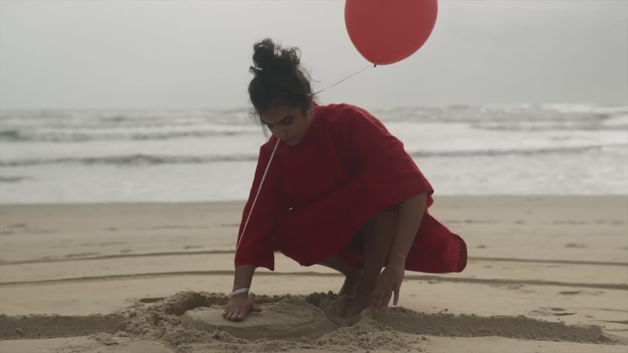 빨간 드레스 를 입은 성인 소녀 가 모래 해변 에 앉아서 낮 에 모래 성 을 형성 하는 모래 를 가지고 놀고 있다