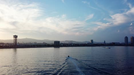 Pull-in-aerial-shot-of-a-boat-entering-the-coast-of-Barcelona-during-a-beautiful-sunset-with-views-over-the-city-skyline