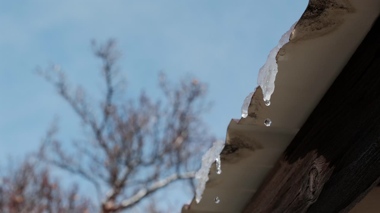 Un primo piano cinematografico al rallentatore cattura l'acqua che gocciola da una piattaforma di ghiaccio che si scioglie su un tetto. Le goccioline illuminate dal sole cadono su un cielo invernale sfocato, illustrando il processo naturale di un disgelo invernale.