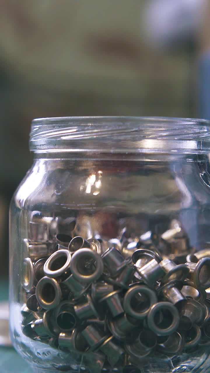 CU: Different silver eyelet parts lie in transparent glass jar against blurry press machine in workshop close view