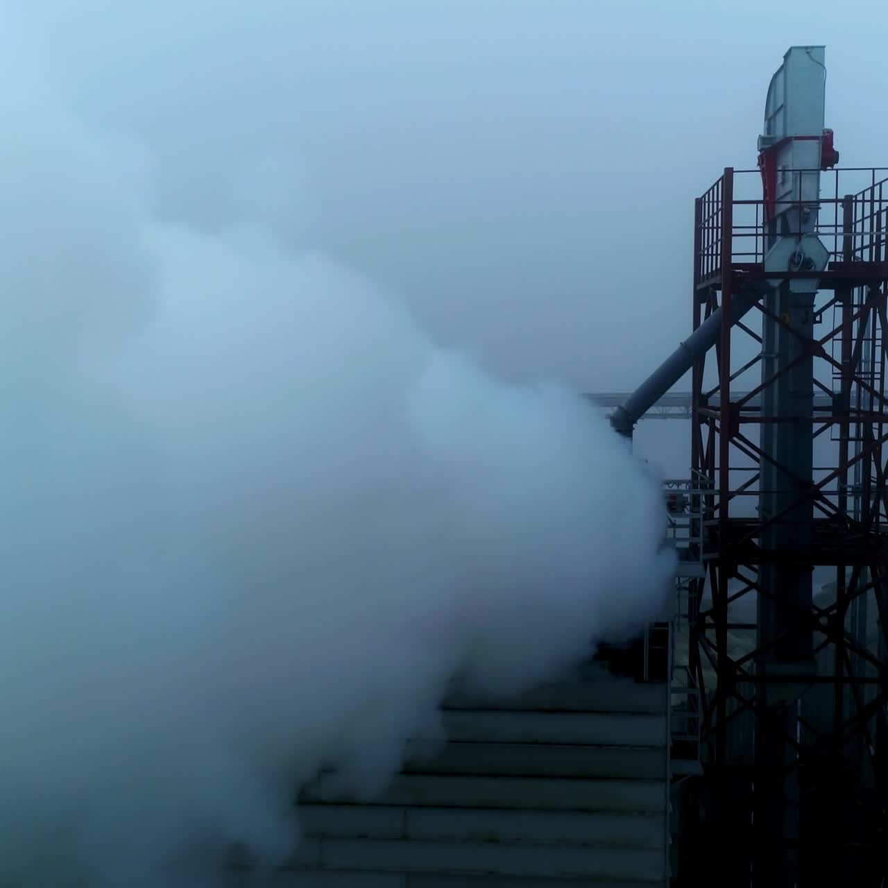 High metal constructions, supports and pipes at the modern elevator plant. Thick white smoke coming into camera. Grey foggy day backdrop
