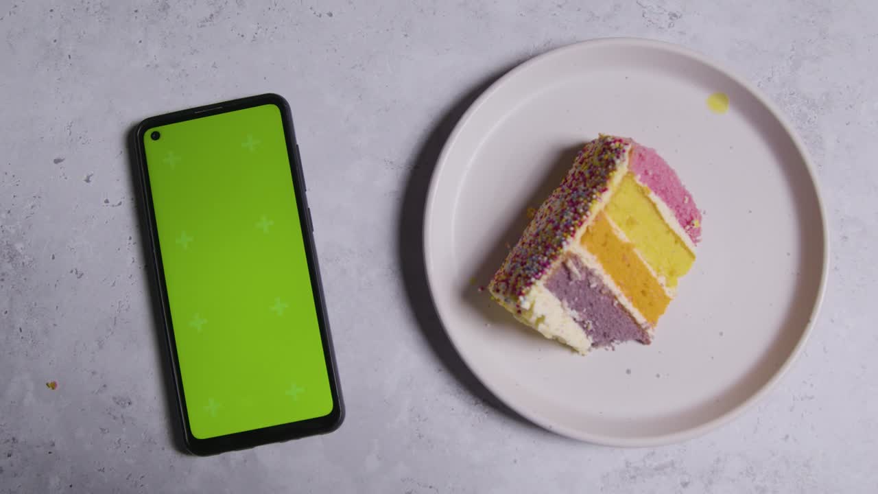 fotografía cenital de un teléfono móvil de pantalla verde con una rebanada cortada del pastel de celebración del arco iris en el plato