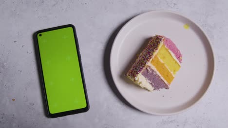 Fotografía-Cenital-De-Un-Teléfono-Móvil-De-Pantalla-Verde-Con-Una-Rebanada-Cortada-Del-Pastel-De-Celebración-Del-Arco-Iris-En-El-Plato